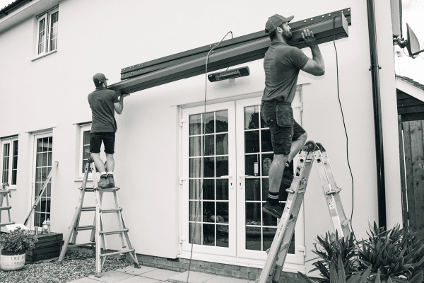 Two men on ladders install an awning above a patio door on a white house. The scene includes plants and paving stones.