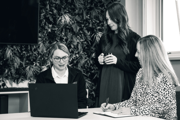 Three women are engaged in discussion around a laptop in an office setting. One woman types, another takes notes, and the third holds a mug, with a plant wall in the background.