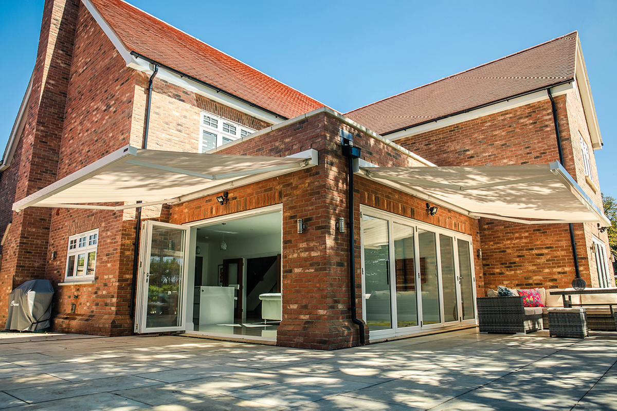 Retractable awnings extend over a patio, attached to a brick house with large glass doors. Outdoor furniture is arranged on the stone tiles, under a clear blue sky.