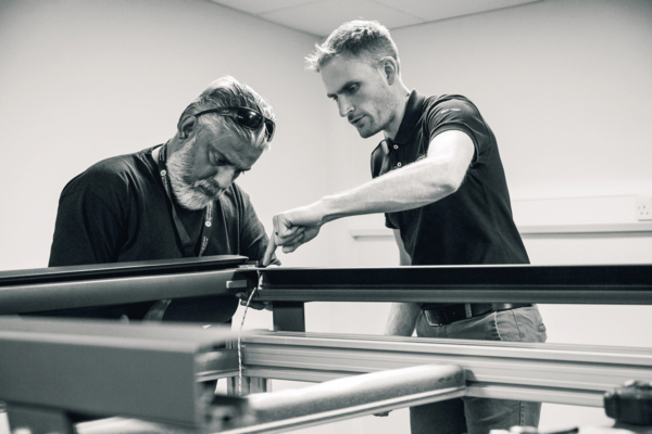 Two men are working together on a large mechanical structure in an indoor setting, focusing intently on making adjustments or repairs to the equipment.