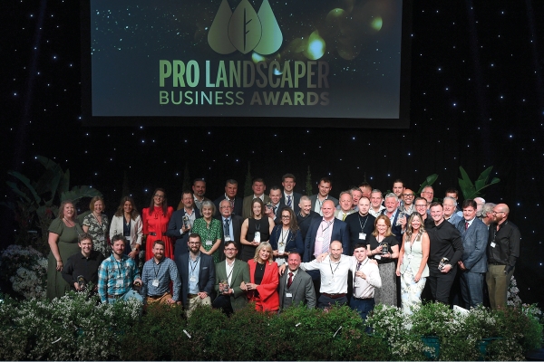 A group of people poses for a photograph on stage, celebrating at an award event. The backdrop displays "PRO LANDSCAPER BUSINESS AWARDS" amidst decorative plants and a starry background.