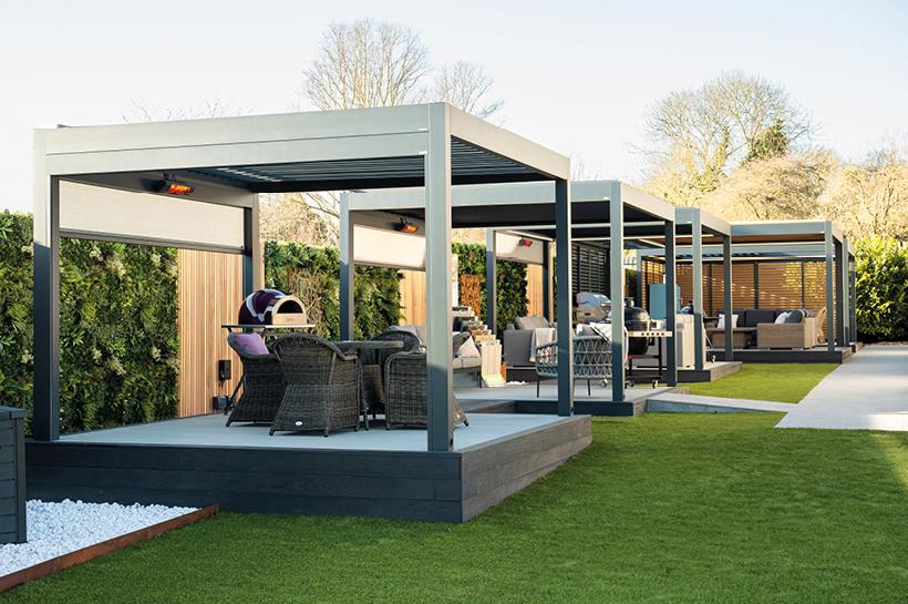 Modern outdoor seating areas sit beneath metal pergolas, featuring wicker chairs and tables. Surrounded by greenery and wooden panels, they occupy a neatly landscaped garden with vibrant grass and trees.