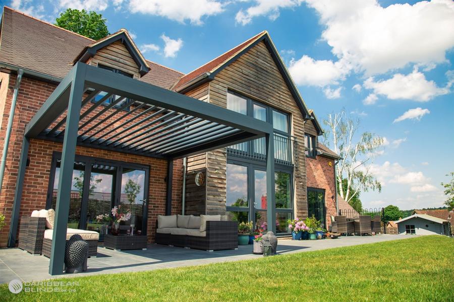 A modern pergola covers a patio with wicker furniture adjacent to a brick house. Lush greenery surrounds the area, and a clear, sunny sky brightens the scene