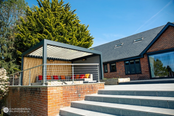 A modern patio gazebo, with colorful seating, stands on a brick platform by a residential building. Situated near a large tree, it overlooks a wide stone staircase. "Caribbean Blinds" logo visible.
