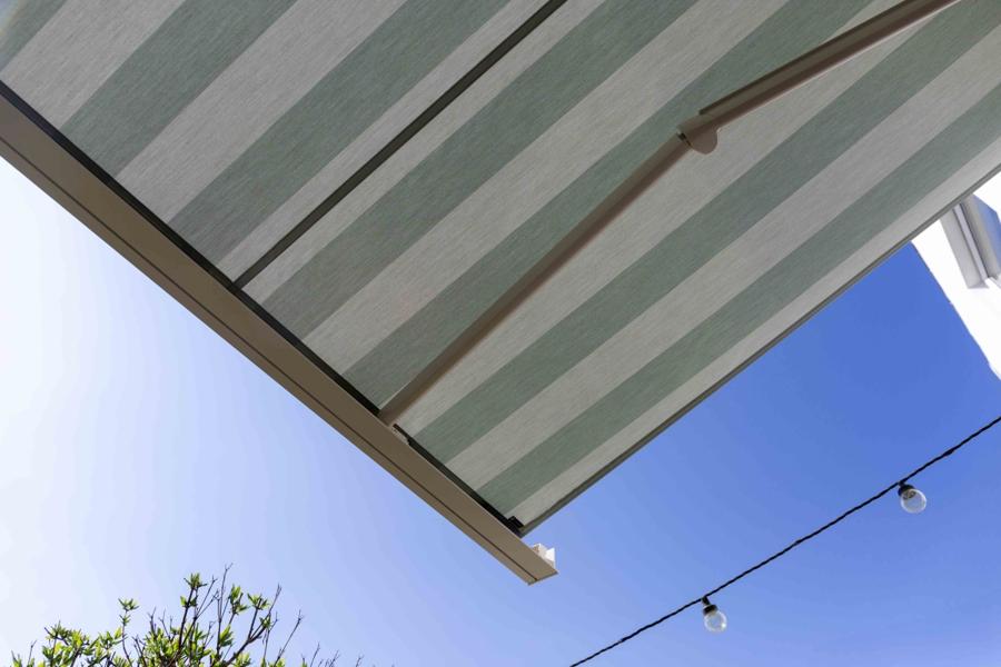 A striped retractable awning shades an outdoor area, partially blocking sunlight. Above, a clear blue sky is visible, while a string of light bulbs hangs nearby, suggesting a relaxing outdoor setting.