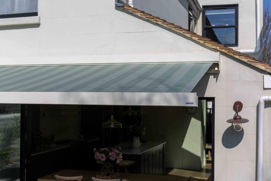 A retractable awning extends from the exterior of a house, providing shade. The surrounding context features a modern kitchen visible through an open door, with decor including a table and flowers.