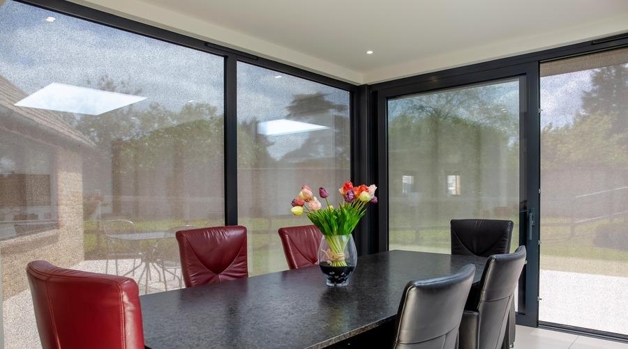 A black dining table holds a vase with colorful tulips, surrounded by red and black chairs. Large windows reveal an outdoor patio with trees and furniture visible in sunlight.