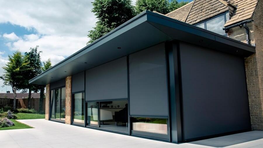 A modern building features large, dark retractable external blinds on its glass facade, set against a manicured lawn and under a partly cloudy sky with nearby greenery. 