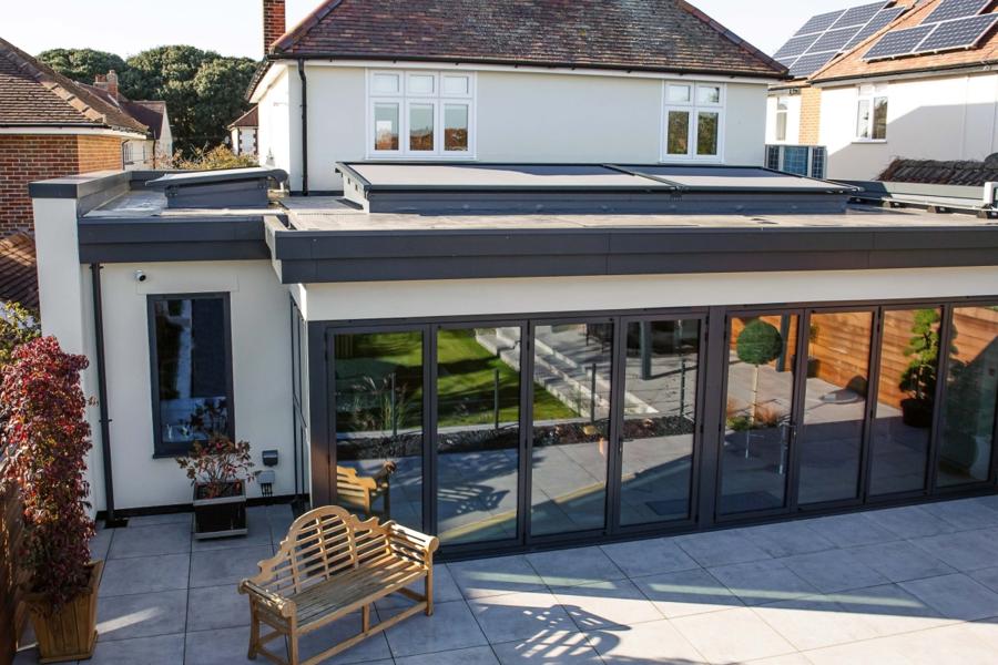 A modern house features a flat roof and large glass doors, with a wooden bench on a tiled patio. It is surrounded by other houses, some with solar panels.