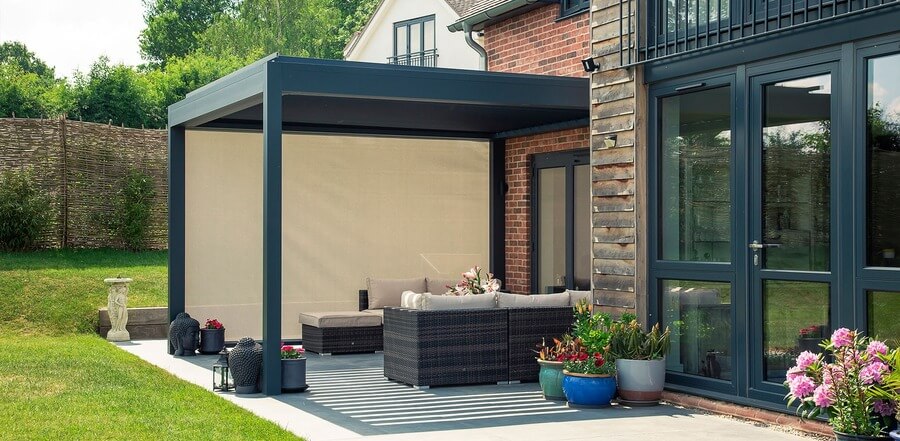 A modern pergola shelters wicker furniture with cushions, decorated with potted plants. It is situated on a patio adjacent to a brick house, with a grassy lawn and wooden fence.