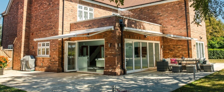 A brick house features open sliding glass doors leading to a patio. Outdoor furniture is arranged nearby on the tiled surface, situated in a sunny backyard with grass and trees.