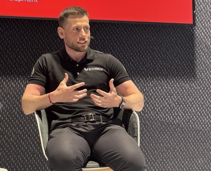 Stuart in a black polo shirt is gesturing while speaking on a stage with a dark, textured backdrop. 