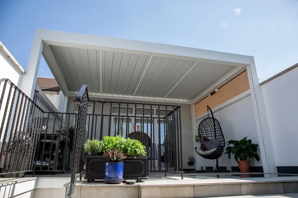 Modern garden terrace with a white louvred outdoor living pod, black railings, hanging chair and potted plants.