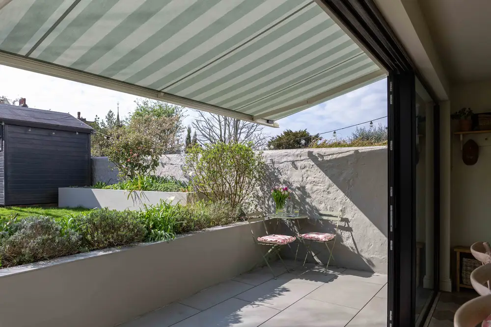 Small patio with a striped retractable awning, bistro table and chairs, overlooking a walled garden.